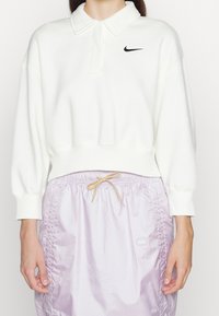 Sudadera blanca con cuello de solapa, puños elásticos y un logo negro de Nike. Combinada con pantalones cortos lavanda ligeros con cordón ajustable.