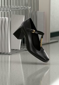 Μαύρο δερμάτινο τακούνι Mary Jane με λεπτομέρεια χρυσής αλυσίδας, τετράγωνη μύτη και χοντρό τακούνι, τοποθετημένο όρθιο σε ανακλαστική επιφάνεια κοντά σε μεταλλικό οργανωτή αρχείων.