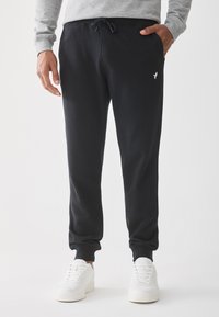 Navy sweatpants featuring a drawstring waist, side pockets, and a small white logo near the hip, paired with white sneakers.