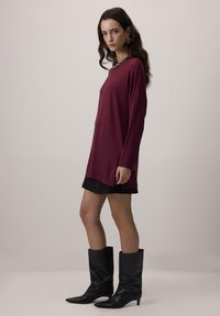 Sieviete, ģērbusies garām piedurknēm bordo topā, melnā mini svārku, melnās vidus teļu zābakos, ar sudraba ķēdes kaklarotu un auskariem aplī, stāv sānis.