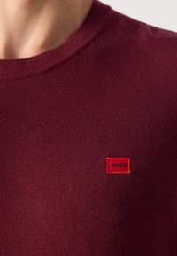 Maroon pleteni pulover s okruglim izrezom i malim crvenim pravokutnim oznakom s natpisom "HUGO" na gornjem dijelu prsnog koša.
