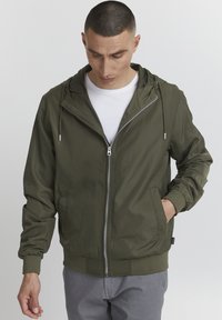 Veste zippée vert olive avec capuche, poignets et taille élastiques. Comprend des poches avant et une texture lisse et légère.
