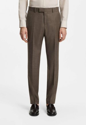 Man wearing brown tailored trousers, black loafers, and a beige long-sleeve shirt, standing against a plain white background.