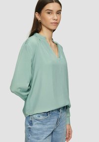 Mintgrüne Bluse mit langen Ärmeln und V-Ausschnitt aus weichem, leichtem Stoff, mit gerafften Schulterakzenten und elastischen Bündchen, kombiniert mit hellblauen Jeans.