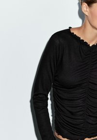Top nero a maniche lunghe con dettagli arricciati, scollatura a onde e design aderente. Realizzato in un tessuto leggero con una texture morbida.
