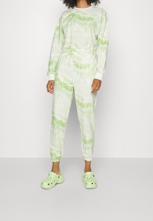 Person wearing a green and white tie-dye sweatshirt and joggers set with light green clogs, standing against a plain light background.