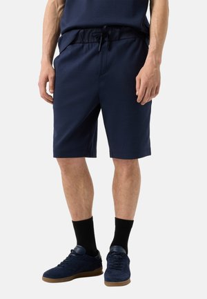 Mand iført marineblå shorts med snøre, sorte crew-sokker og mørkeblå sneakers på en ensfarvet lys baggrund.