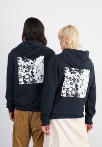 Tumesinine kapuutsiga pusakas, mille seljal on valge abstraktne muster, ning millel on känguru-tasku ja nööriga kapuuts. Pehme puuvilla segu materjal.