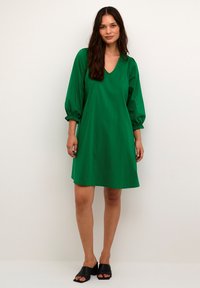 Grön kort klänning med V-ringning, puffärmar som samlas vid ärmslutet och en lös passform. Bärs med svarta slidesandaler. Slät textur.