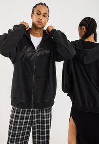 Two models wearing oversized black zip-up hoodies, one facing forward adjusting the hood, the other turned back with braided hair.