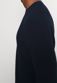 Suéter de punto azul marino con cuello redondo y mangas largas, que muestra un patrón texturizado y una silueta ajustada desde la vista lateral.