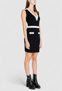 Schwarzes ärmelloses Kleid mit V-Ausschnitt, weißen Akzenten an Schultern und Taille sowie zwei vorderen Taschen mit Knöpfen. Kombination mit schwarzen Ankle Boots.