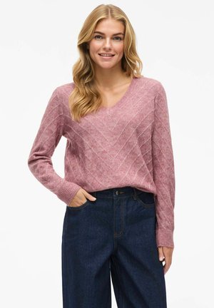 Pull rose en V avec un motif en chevrons texturé, associé à un jean à taille haute en denim foncé. Manches longues avec des poignets côtelés.