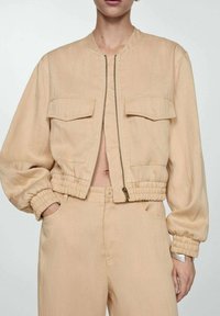 Beige cropped bomberjack met ritssluiting, twee voorzakken en elastische manchetten en zoom. Gemaakt van lichte stof met een gladde textuur.