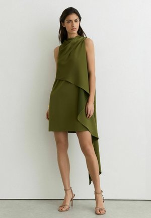 Žena stojaca oblečená v asymetrických bezrukávových olivovo zelených šatách s nariasenou látkou a telovými sandálmi na vysokom opätku so remienkami.