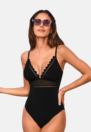 Bañador de una pieza negro con canalé, bordes festoneados, inserciones de malla transparente y piernas de corte alto, con tirantes delgados ajustables.