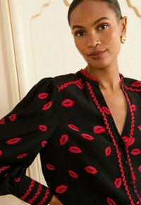 Blusa nera con motivi di labbra rosse, scollatura a scalloped e maniche a palloncino elasticizzate con bordi rossi. Il tessuto testurizzato aggiunge profondità.