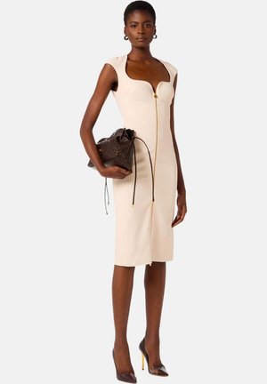 Donna che indossa un vestito beige aderente senza maniche con cerniera frontale, tiene una borsa scura marrone testurizzata con coulisse e indossa tacchi alti neri.