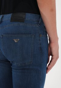 Jeans din denim albastru închis, cu un buzunar ce are un logo argintiu, etichetă din piele pe talie și detalii slim fit pe spate.