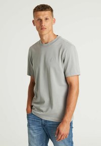 Siva pamučna majica s kratkim rukavima, okruglim izrezom i suptilnim teksturiranim uzorkom. Ima mali, utisnuti logo na prsima. Uparena je s plavim trapericama.