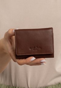 Bruine leren portemonnee met een gestructureerde afwerking, opgevouwen ontwerp en geperst logo; vastgehouden door een hand met lichte nagellak.
