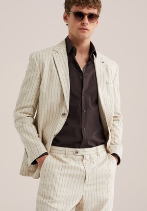 Mand iført cremefarvet jakkesæt med fine striber, sort skjorte med knapper og solbriller, hænderne i lommerne, foran en ensfarvet beige baggrund.
