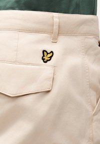 Poche de pantalon beige avec rabat et petit logo oiseau jaune brodé au-dessus de la poche, porté avec une chemise vert foncé.