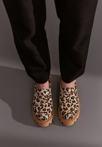 Schwarz-gelb gefleckte Slipper mit einer klobigen tanfarbigen Sohle, ausgestattet mit einem weichen Stoffoberteil und einem breiten Riemen über dem Spann, kombiniert mit schwarzen Hosen.