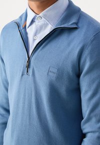 Pull-over en maille bleu clair avec une fermeture éclair au quart, présentant une finition texturée, un patch logo ton sur ton, et un col superposé sur une chemise à carreaux.