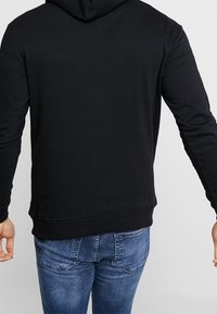 Schwarzer Hoodie mit Kängurutasche aus weichem Stoff, mit gerippten Bündchen und Bund, von hinten gesehen, kombiniert mit blauen Jeans.