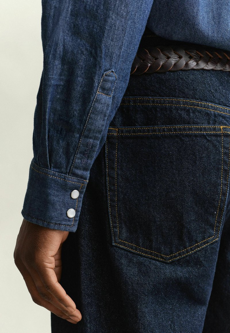 Close-up of a person wearing dark blue denim shirt with buttoned cuff and dark blue jeans with yellow stitching and a braided brown leather belt.