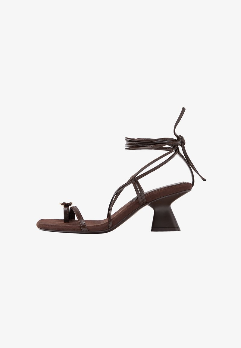 Braune Sandale mit Riemchen, quadratischer Zehenpartie, niedrigem ausgestelltem Absatz, Zehenring und Schnürung um den Knöchel vor weißem Hintergrund.