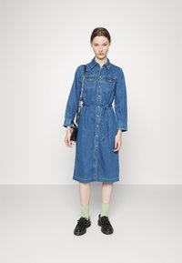 Vestido de camisa de ganga azul, apresentando mangas longas, fecho frontal com botões, dois bolsos no peito, cintura cingida por um cinto e bainha reta. Sapatos pretos e meias verdes.