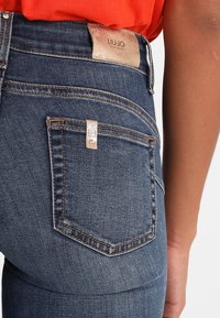Jeans de denim azul oscuro con costuras doradas, que presentan un prominente bolsillo trasero y un parche de logo de cuero texturizado en la cinturilla.