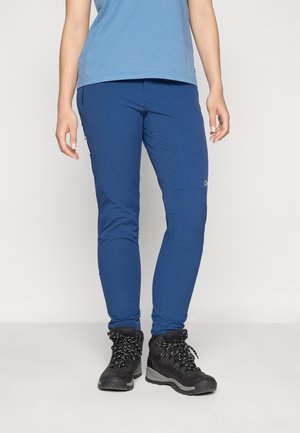 Person iført blå slim-fit outdoorbukser og sorte vandrestøvler, stående mod en ensfarvet lys baggrund.