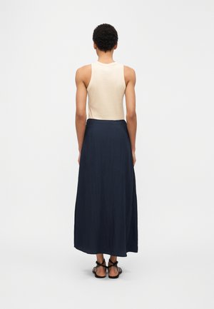 Person mit kurzen lockigen Haaren, die ein ärmelloses beigefarbenes Oberteil, einen langen dunkelblauen Rock und schwarze Sandalen mit Riemchen trägt, steht mit dem Rücken zur weißen Hintergrundwand.