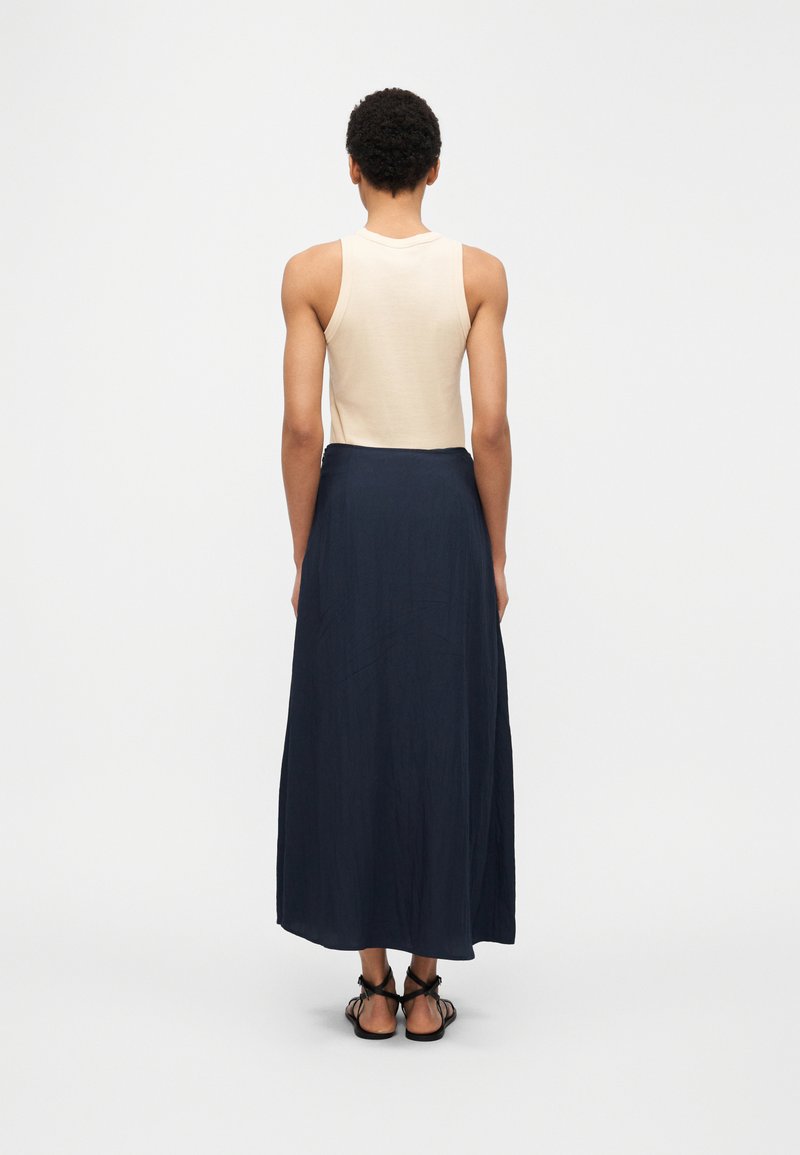 Άτομο με κοντά σγουρά μαλλιά που φοράει αμάνικο μπεζ τοπ, μακριά ναυτικά φούστα και μαύρα σανδάλια με λουριά, στέκεται με την πλάτη γυρισμένη σε λευκό φόντο.