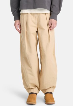 Beżowe spodnie z szerokimi nogawkami, plisami i elastycznymi ściągaczami, wykonane z lekkiego materiału. W połączeniu z ciemnoszarym swetrem i brązowym obuwiem.