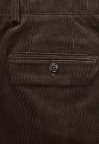 Brauner Cordstoff mit vertikaler Rippung, ausgestattet mit einer Rückentasche, die durch einen schwarzen Knopf gesichert ist. Glatte Textur und detaillierte Nähte.