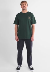 T-shirt en coton vert foncé avec un petit motif brodé au centre. Porté avec un pantalon foncé et des chaussures noires. Col rond simple.