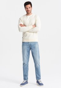 Sweatshirt crème à encolure ronde, avec des poignets et un ourlet côtelés, associé à un jean bleu clair et des baskets bleu foncé. Design simple, texture lisse.