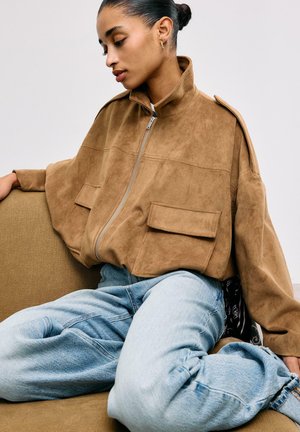 Žena sedící na hnědé pohovce, oblečená v béžové semišové bundě na zip s velkými kapsami a světle modrých džínách, dívající se dolů s vlasy svázanými do drdolu.