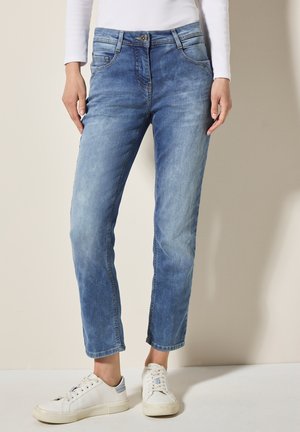 Jeans de denim azul claro con corte recto, cinco bolsillos y cierre de botón; combinados con zapatillas blancas.