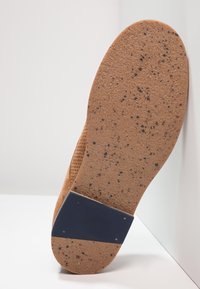 Gumová podešev v světle hnědé barvě s tmavými skvrnami. Obsahuje strukturovaný povrch a obdélníkový námořnický akcent poblíž základny.