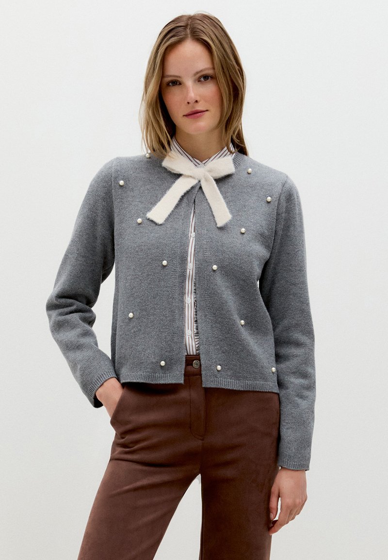 Frau trägt eine graue Strickjacke mit Perlenverzierungen und einer weißen Schleife, über einem gestreiften Hemd und braunen Hosen.