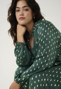 Robe verte avec des motifs de feuilles dorées, présentant un décolleté en V profond et des poignets à volants. Le tissu a une finition douce et texturée avec un tomber fluide.