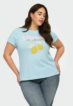 Kvinde med langt mørkt hår iført lyseblå T-shirt med gule citroner og teksten "Limoni di Amalfi", sammen med blå jeans, poserer med hænderne på hofterne.