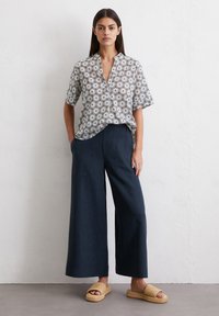 Chemise à manches courtes à motif floral en blanc et marron, associée à un pantalon large bleu marine. Portée avec des sandales à plateforme tissées beiges.