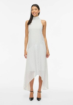 Femme debout portant une robe blanche sans manches à pois noirs et des talons noirs à brides à la cheville sur fond blanc.