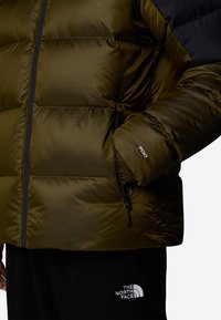 Oliivinvihreä puhvijakku, jossa on tikattu kuvio, mustat yksityiskohdat, vetoketjulliset sivutaskut ja hihan "700" -label. Mustat fleece-housut alla.